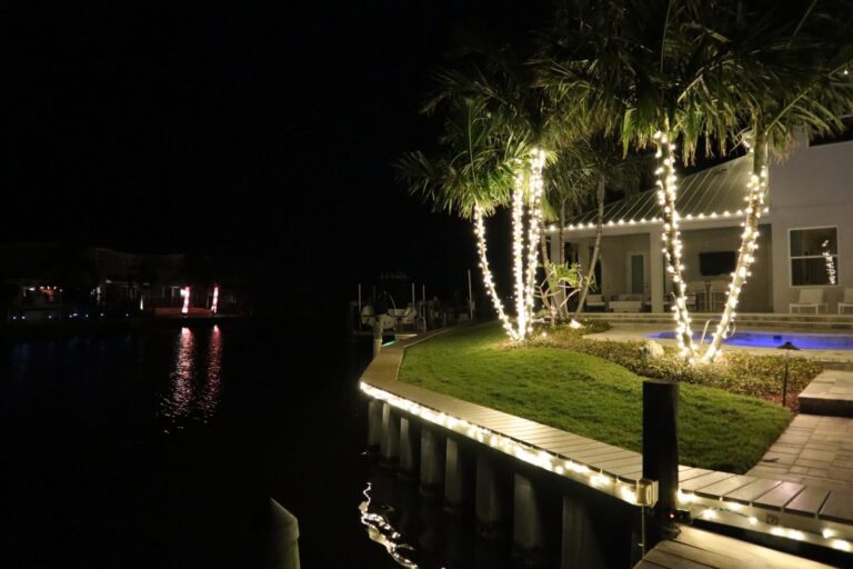 Palm trees and a seawall along the water wrapped in warm white Christmas lights