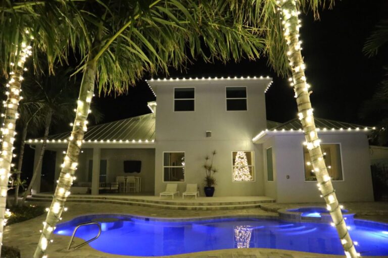 Back of a two-story home lit for Christmas with the pool and palms